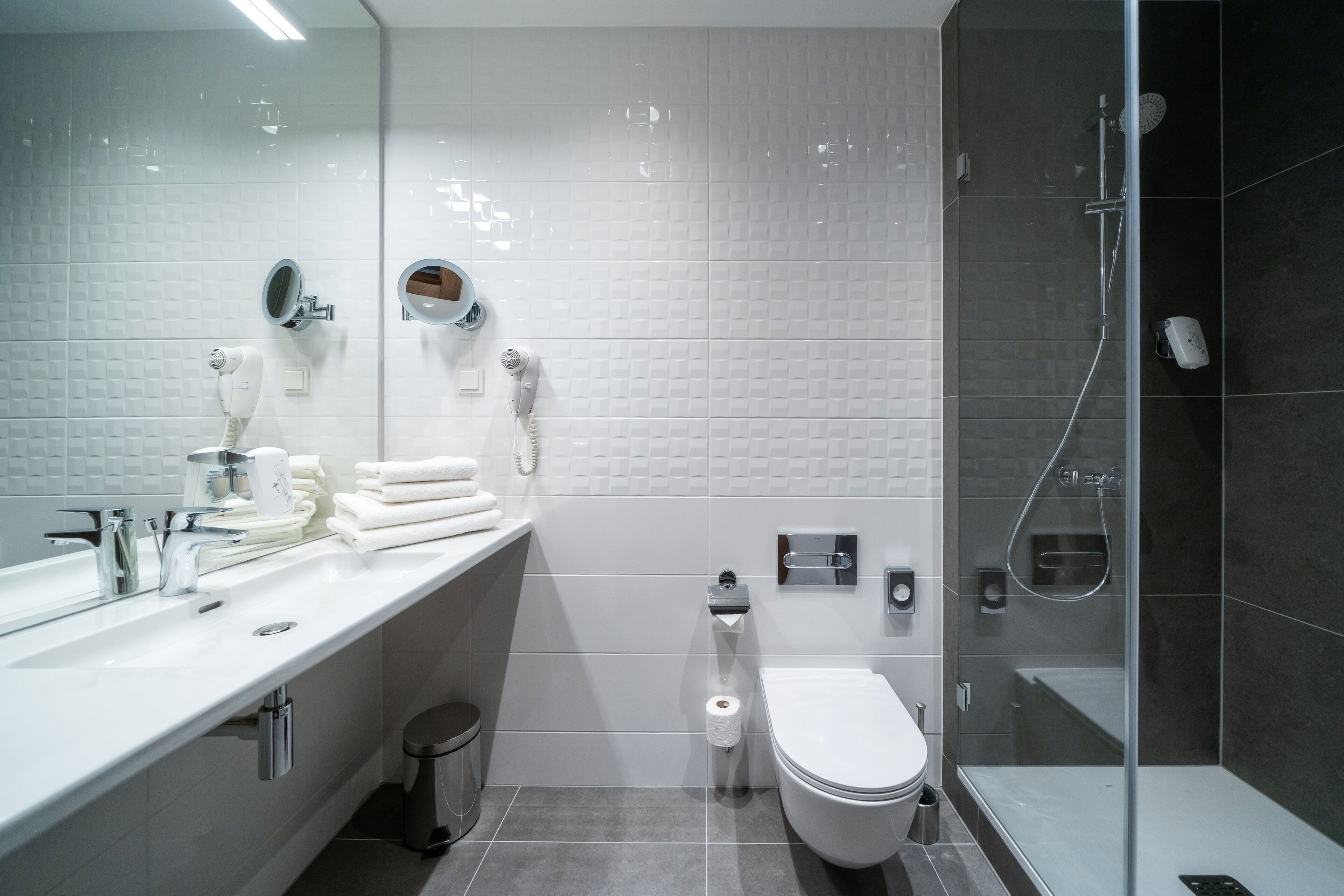 Un baño moderno con lavamanos blanco, inodoro, ducha con mampara de vidrio y toallero.