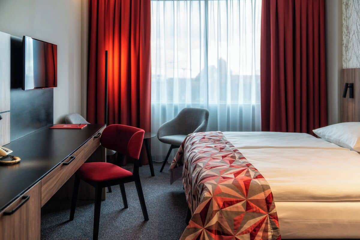 Une chambre d’hôtel comprenant un lit, un bureau avec une chaise, un téléviseur fixé au mur et une fenêtre avec des rideaux.