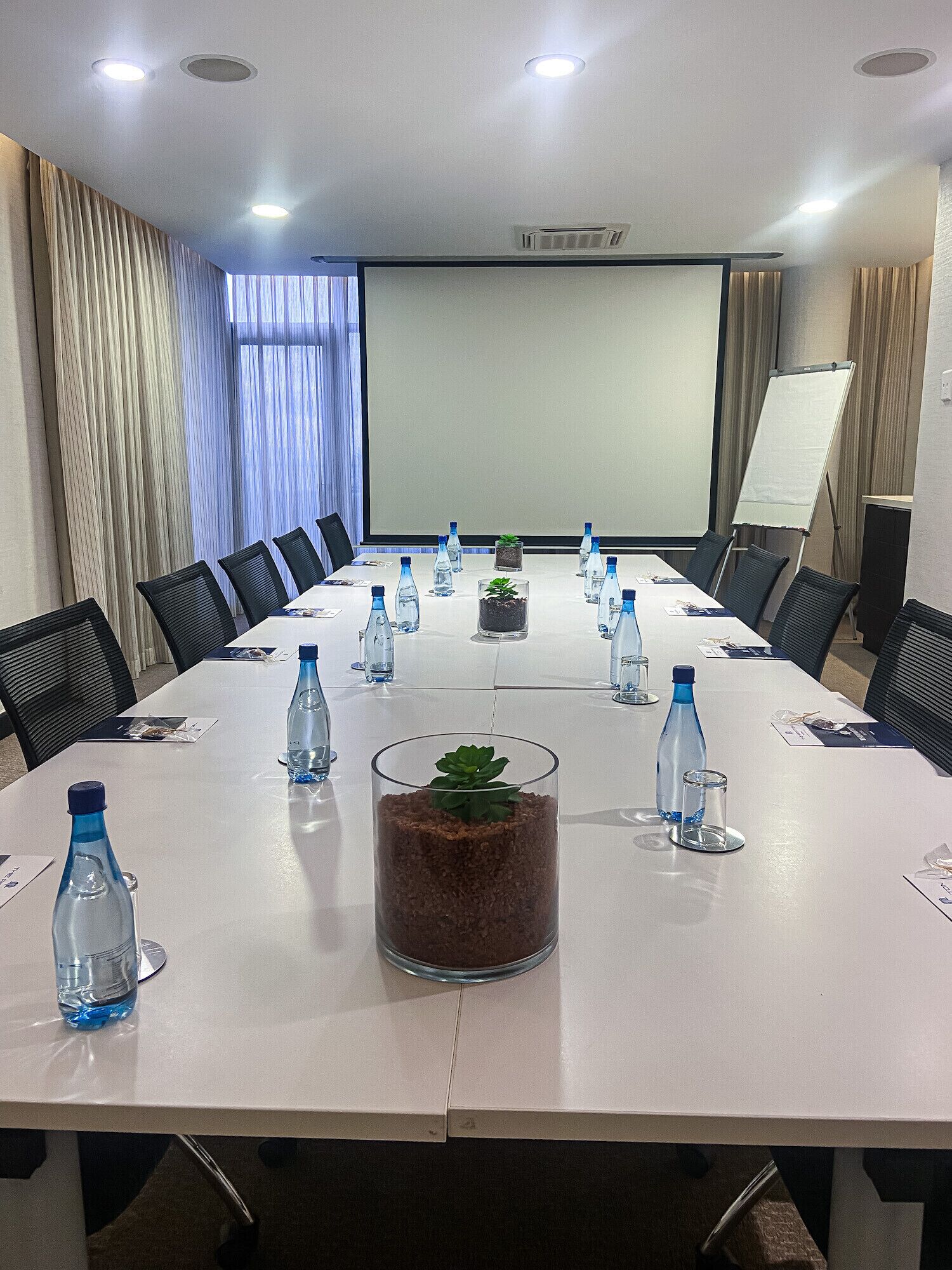 Un salón de conferencias con una larga mesa, sillas, botellas de agua y una pantalla de proyector.