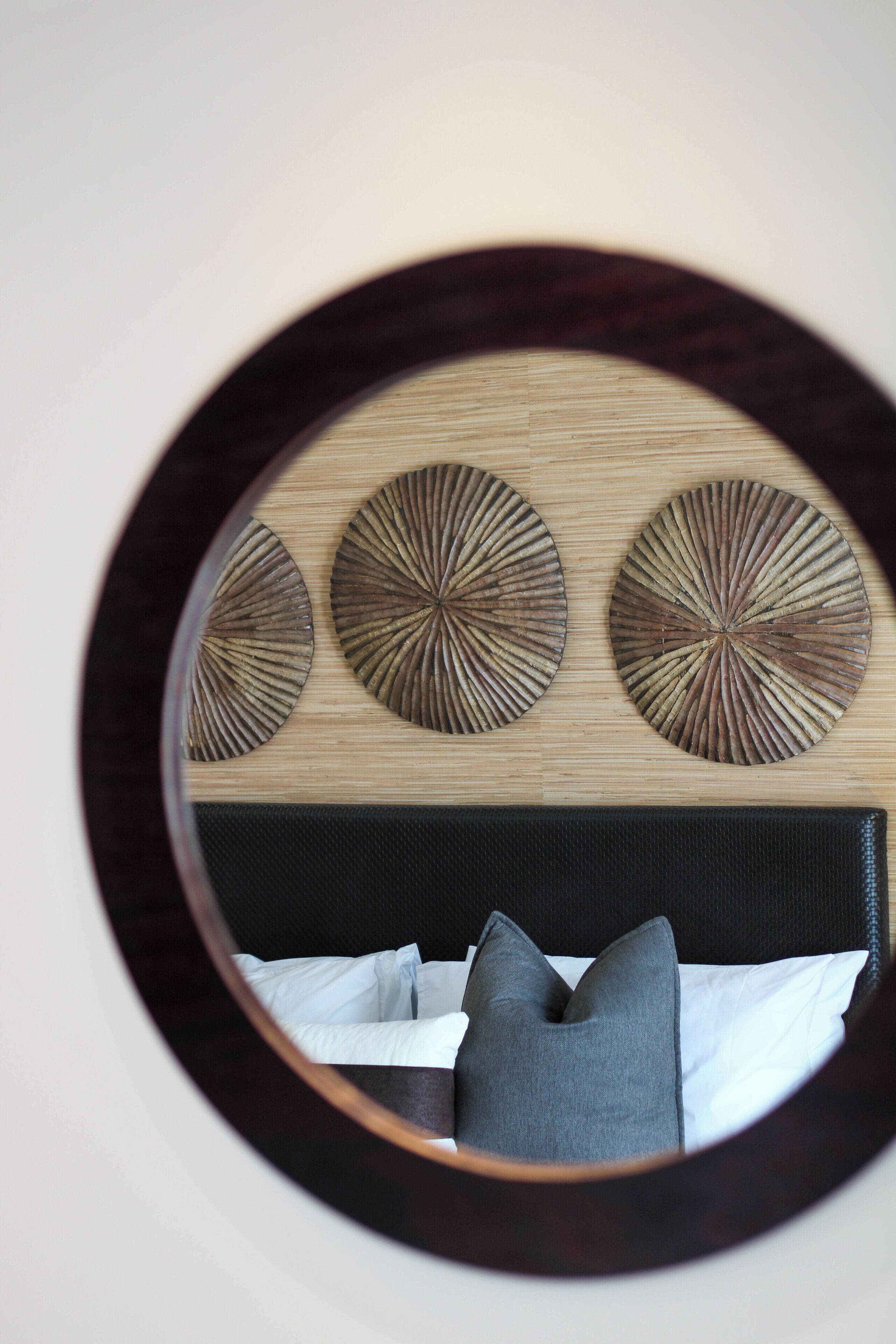 A circular mirror reflecting a bed with a dark headboard, three wall-mounted decorative panels, and two pillows.