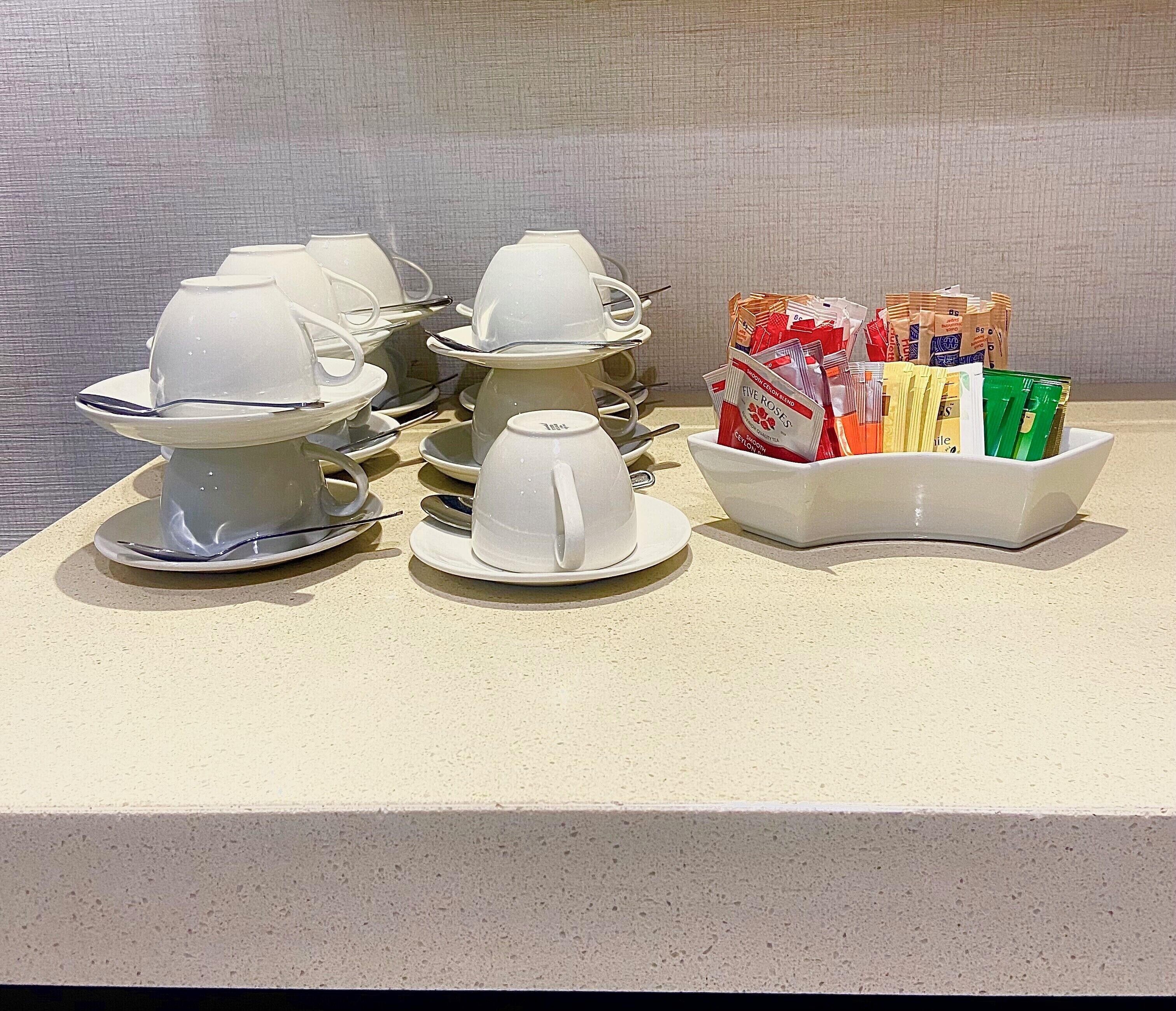 A display of white teacups and saucers, a bowl of tea bags, and a tray of assorted snacks on a countertop.