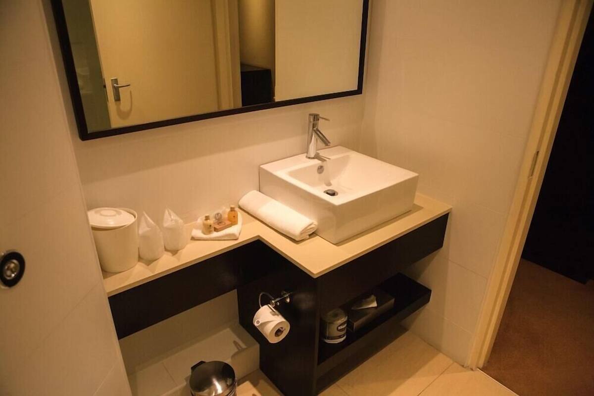 A bathroom with a white sink, a mirror, a towel, toiletries, and a shelf with a roll of toilet paper.