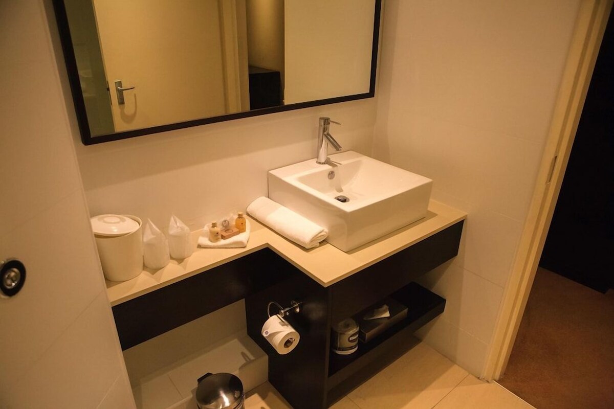 A bathroom with a white sink, a mirror, a towel, toiletries, and a shelf with a roll of toilet paper.