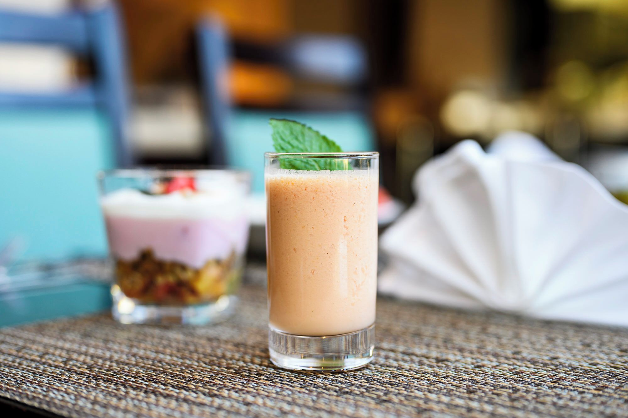 Une boisson servie dans un verre transparent, décorée d’une feuille de menthe, un dessert présenté dans un verre avec des couches apparentes, et une serviette blanche posée sur une nappe à motifs tissés.