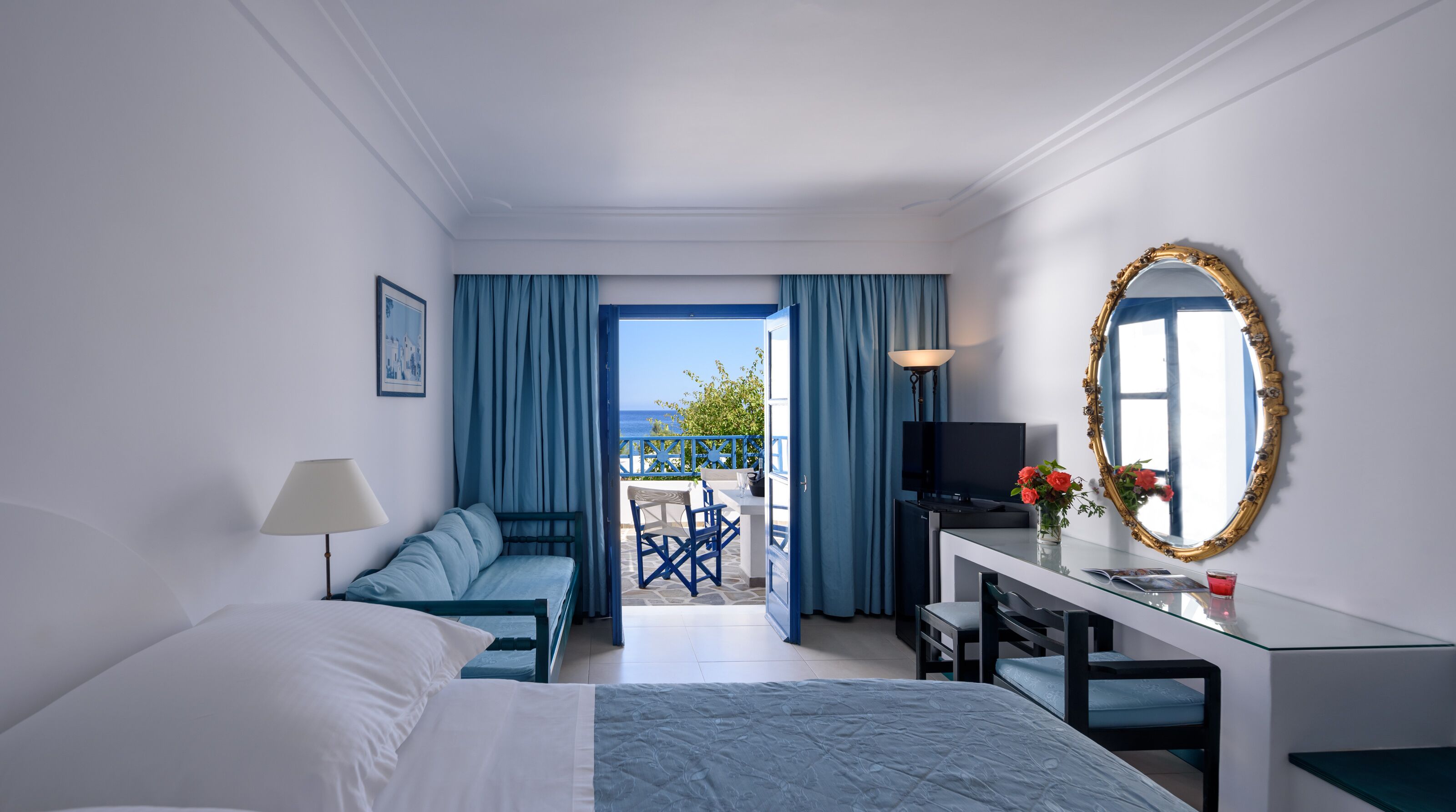 Habitación de hotel con cama, escritorio con televisor, balcón con cortinas azules y un espejo grande y decorativo.