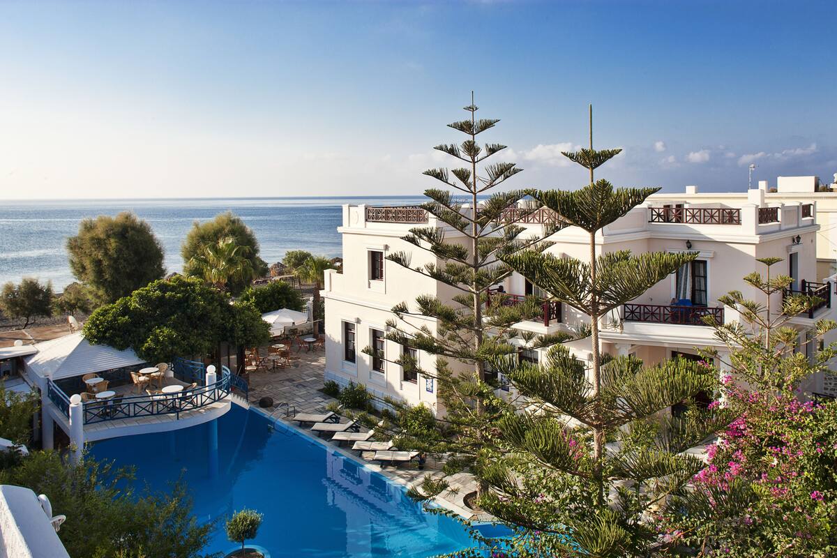 A poolside area with a view of the sea, surrounded by white buildings and lush greenery.