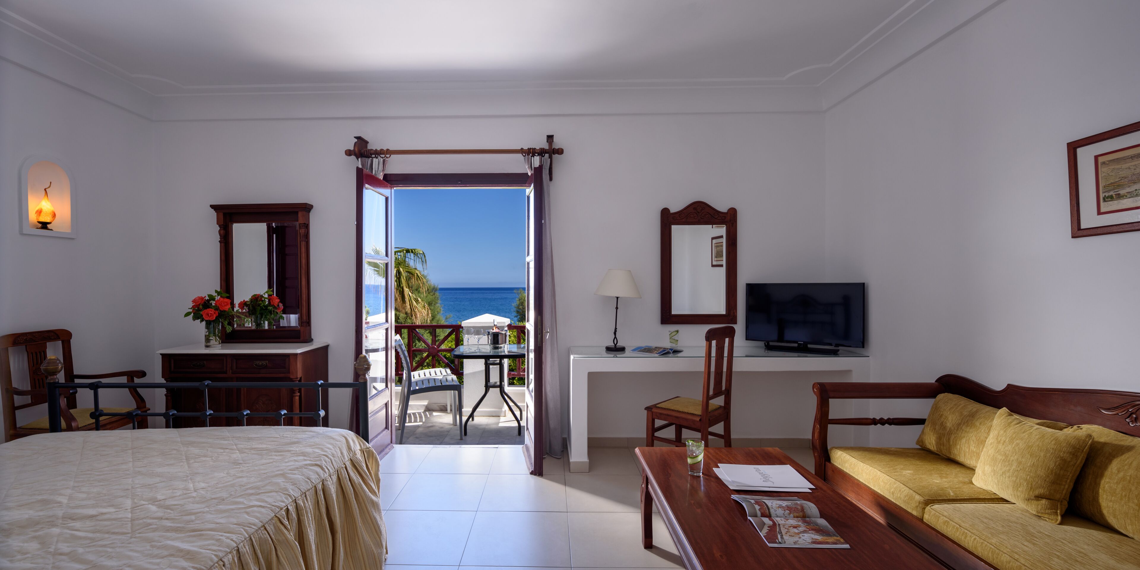 Una camera d'hotel con un letto, un divano, una scrivania con televisore, un balcone vista mare e un piccolo tavolo con lampada.