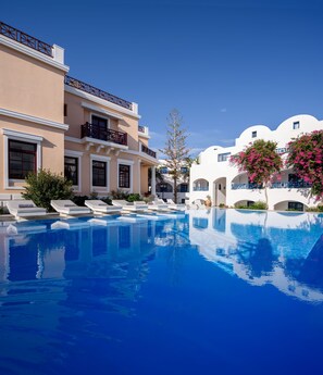 A poolside view of a hotel with a clear blue pool, white and beige buildings, and pink flowering trees. - Veggera Hotel (Santorini)