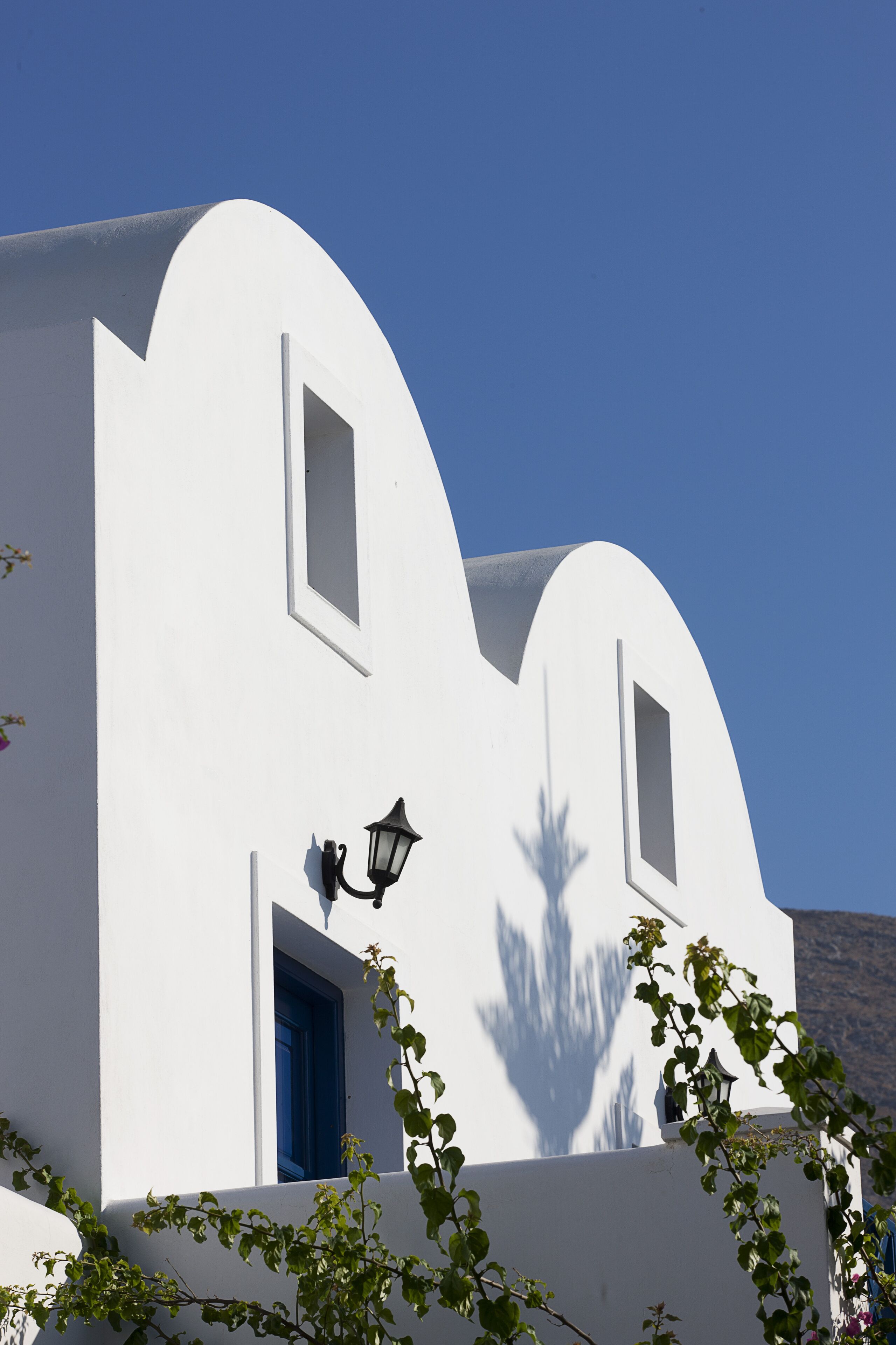 Un bâtiment blanc avec une porte et des fenêtres bleues, une lampe noire fixée au mur et l’ombre d’un arbre.