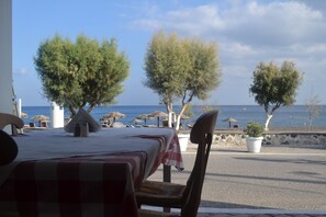 A seaside restaurant with outdoor seating, a view of the sea, and trees. - Veggera Hotel (Santorini)