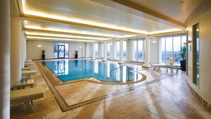 Une piscine intérieure offrant une vue dégagée sur la ville grâce à de grandes fenêtres, un plafond baigné de lumière et un sol carrelé.