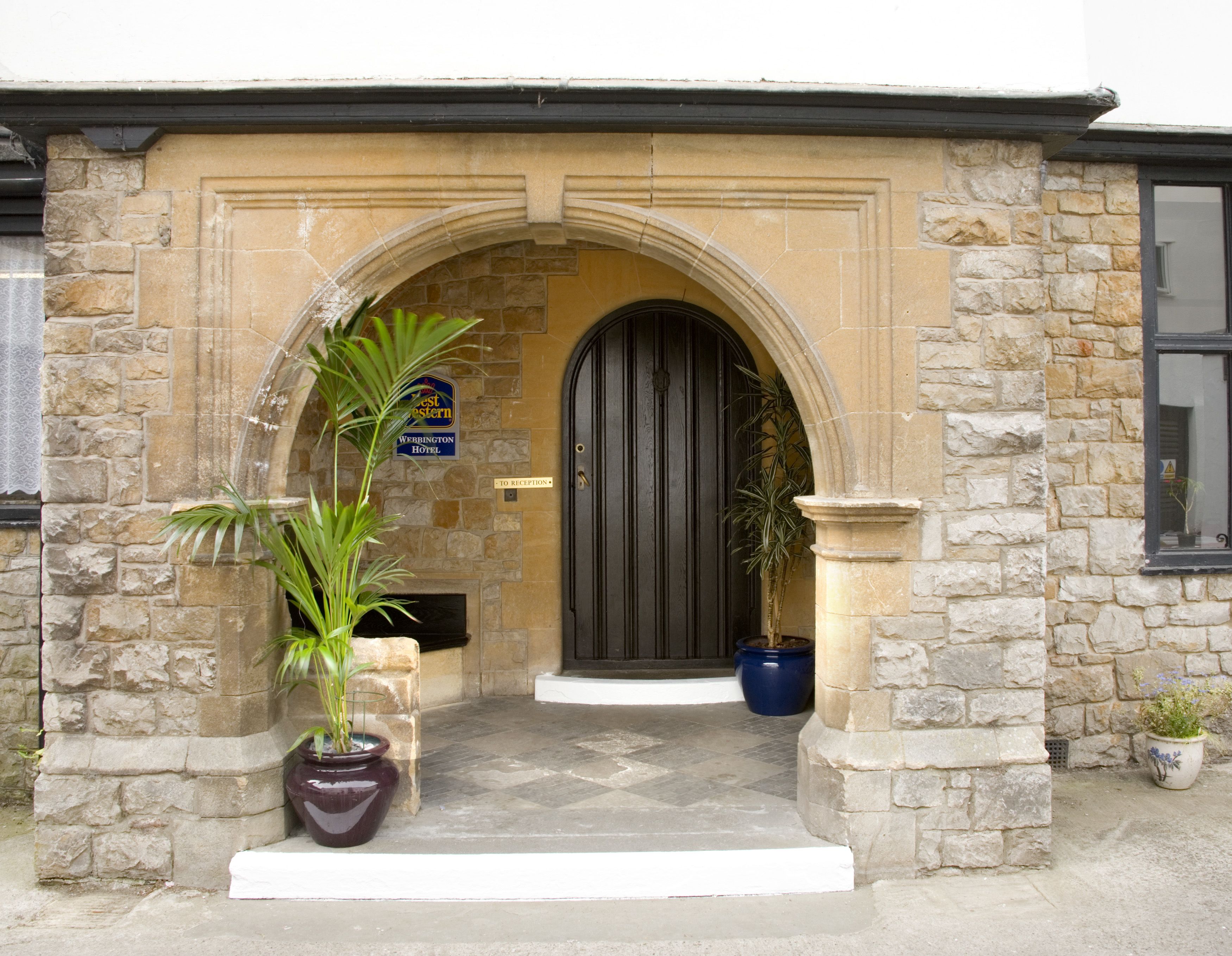 A stone building entrance with a black door, a reception sign, and potted plants.