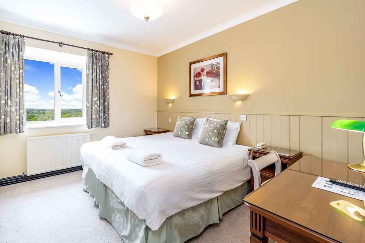 A bedroom with a large bed, two bedside tables, a desk, and a window with curtains.