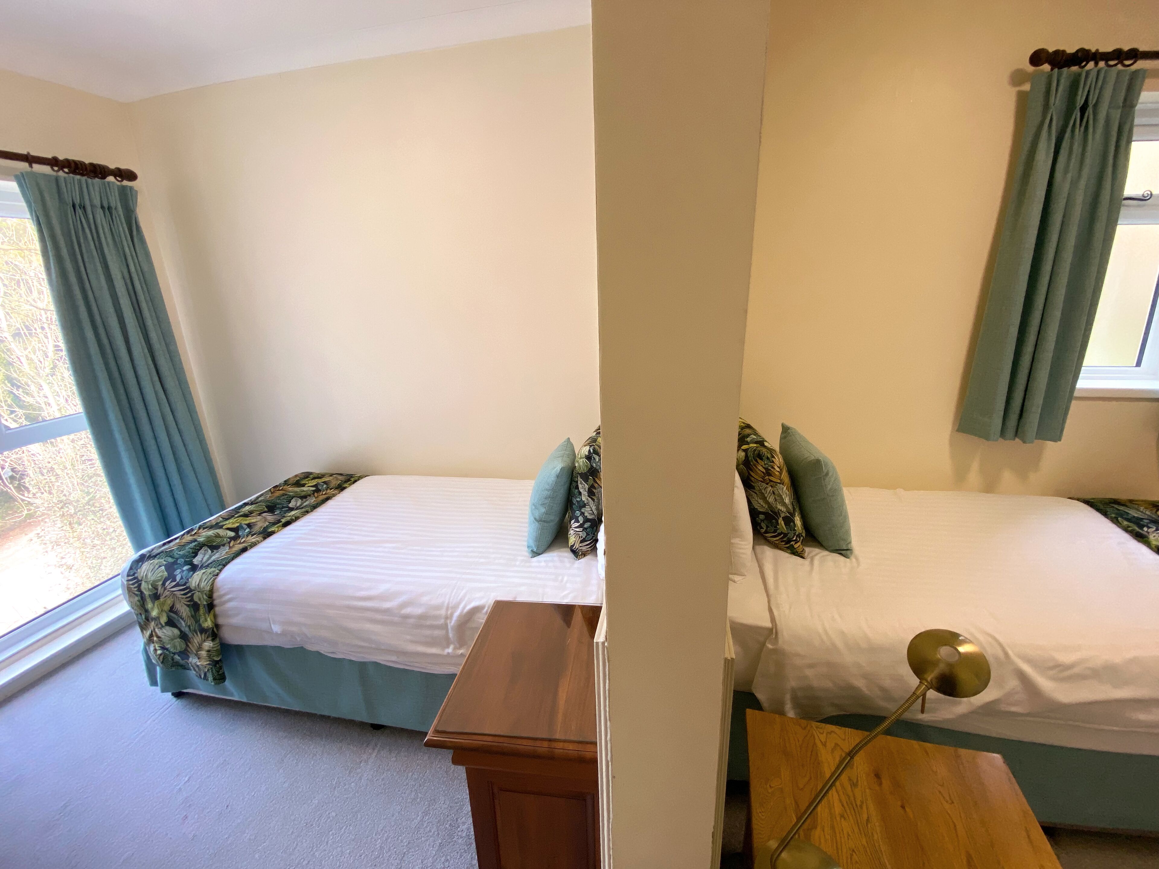 A room with two beds, each with a floral bedspread and matching pillows. There is a wooden nightstand in front of one bed and a lamp on the other. The room has cream-colored walls and a window with blue curtains.