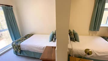 Habitación con dos camas, cada una con una colcha de flores y almohadas a juego. Se observa un mesita de noche de madera frente a una cama y una lámpara sobre la otra. Las paredes son de color crema y hay una ventana con cortinas azules.