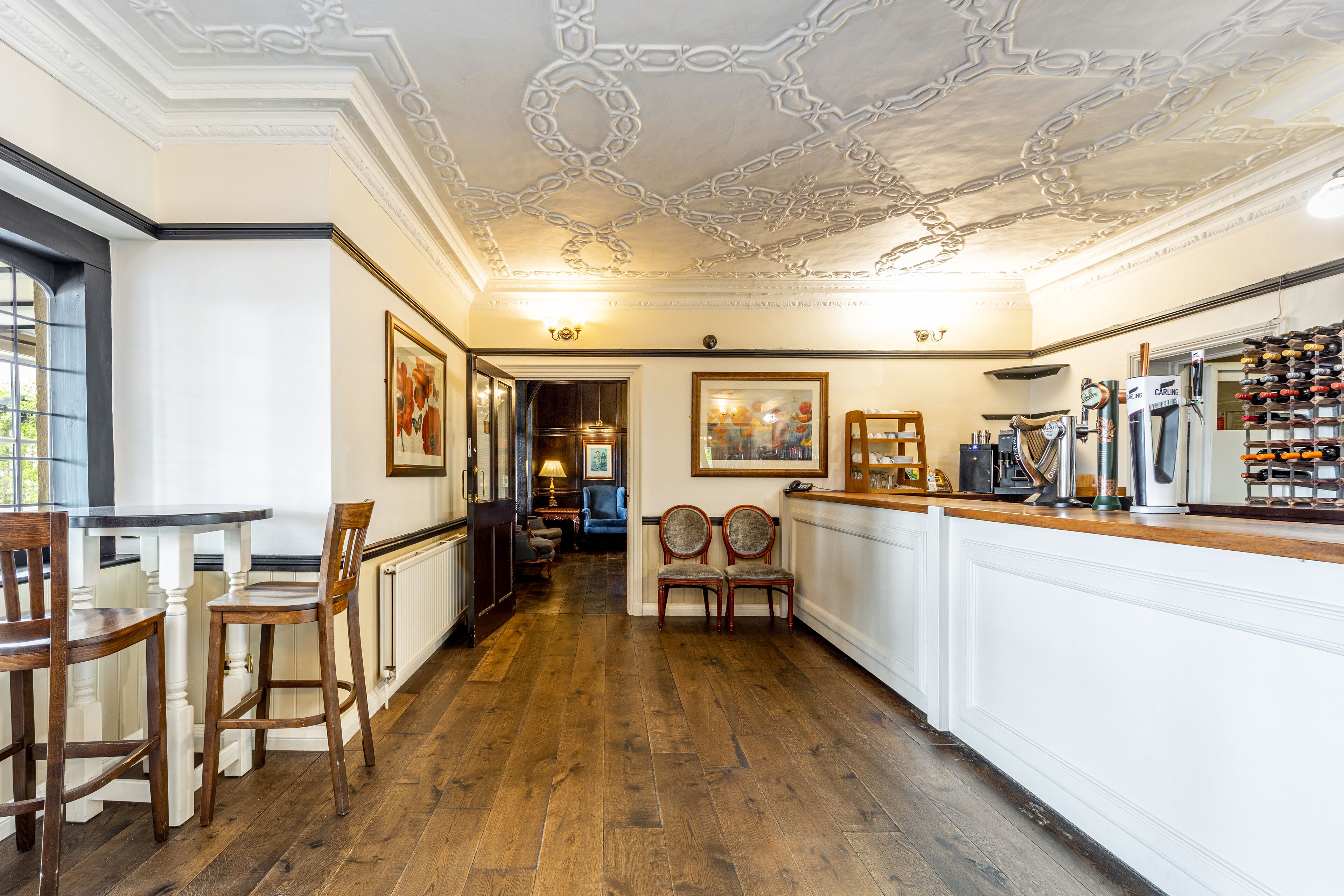 A hotel lobby with a wooden floor, white walls, and a decorative ceiling. There are two wooden chairs and a table near the entrance.