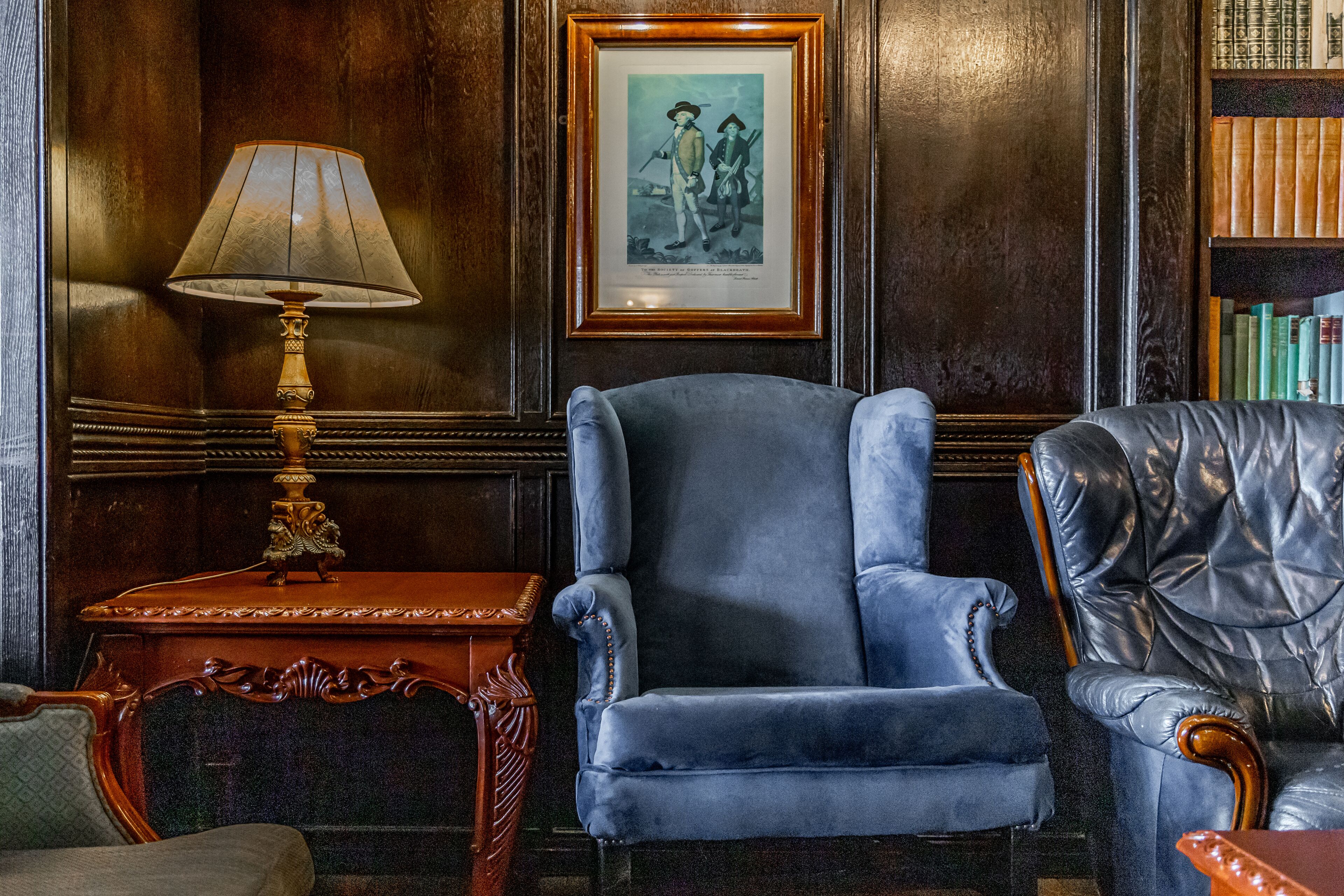 A room with a blue armchair, a wooden side table with a lamp, and a framed picture on the wall.