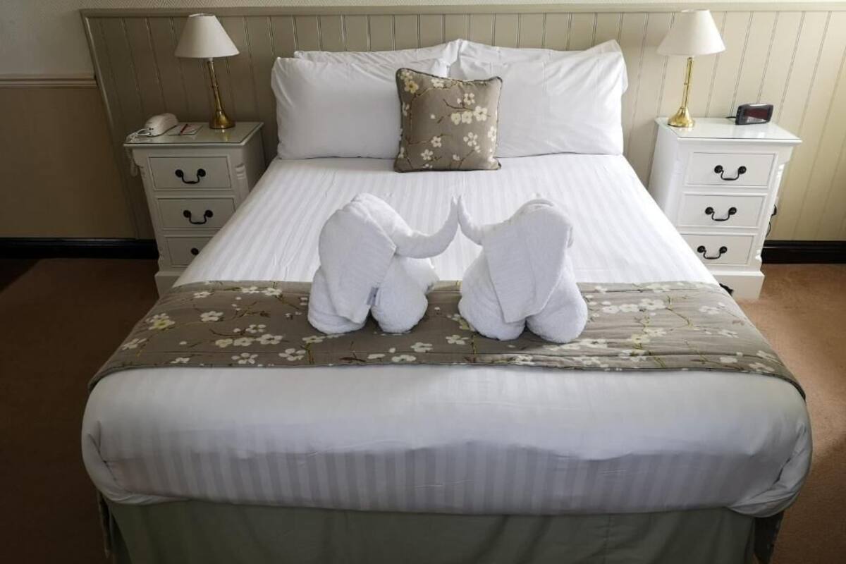 A neatly made bed with white linens, a floral-patterned bedspread, and two towels arranged in a heart shape on the bed.