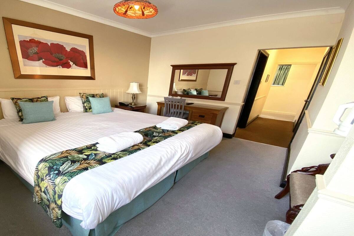 A hotel room with a large bed, a nightstand, a desk with a chair, a mirror, and a door leading to another room.