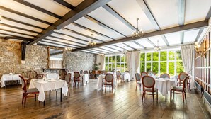 Un vaste espace dédié à la salle à manger, doté de planchers en bois, de tables et de chaises blanches, de grandes fenêtres avec des rideaux et d’un mur en pierre.