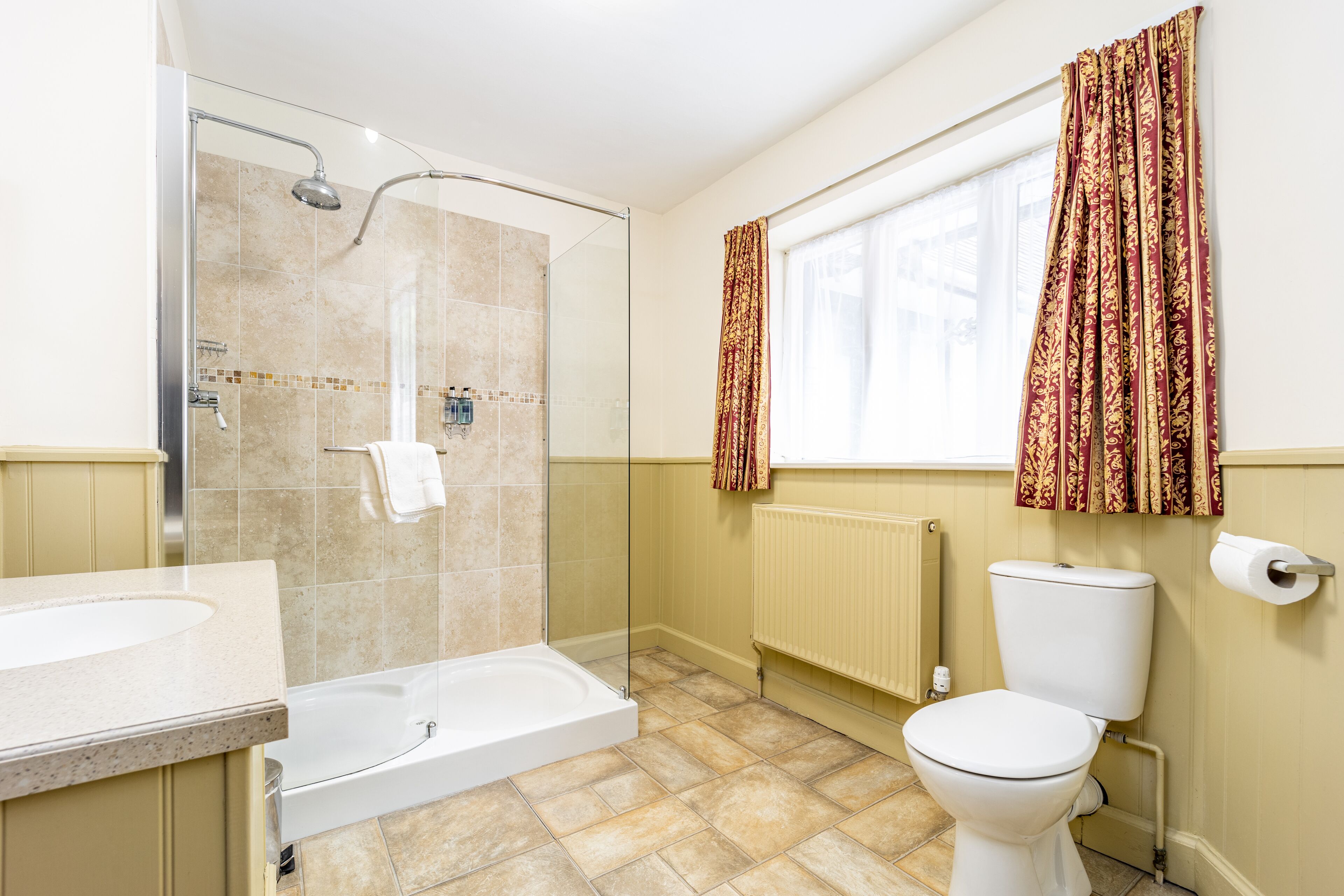 A bathroom with a glass-enclosed shower, a bathtub, a toilet, and a window with patterned curtains.