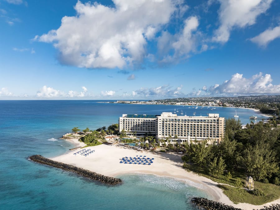 Un hotel frente a la playa con área de piscina, una playa con tumbonas y vistas al océano y al horizonte de la ciudad.