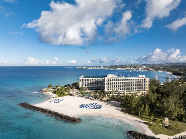 A beachfront hotel with a pool area, a beach with sun loungers, and a view of the ocean and city skyline. - Hilton Barbados Resort (Bridgetown)
