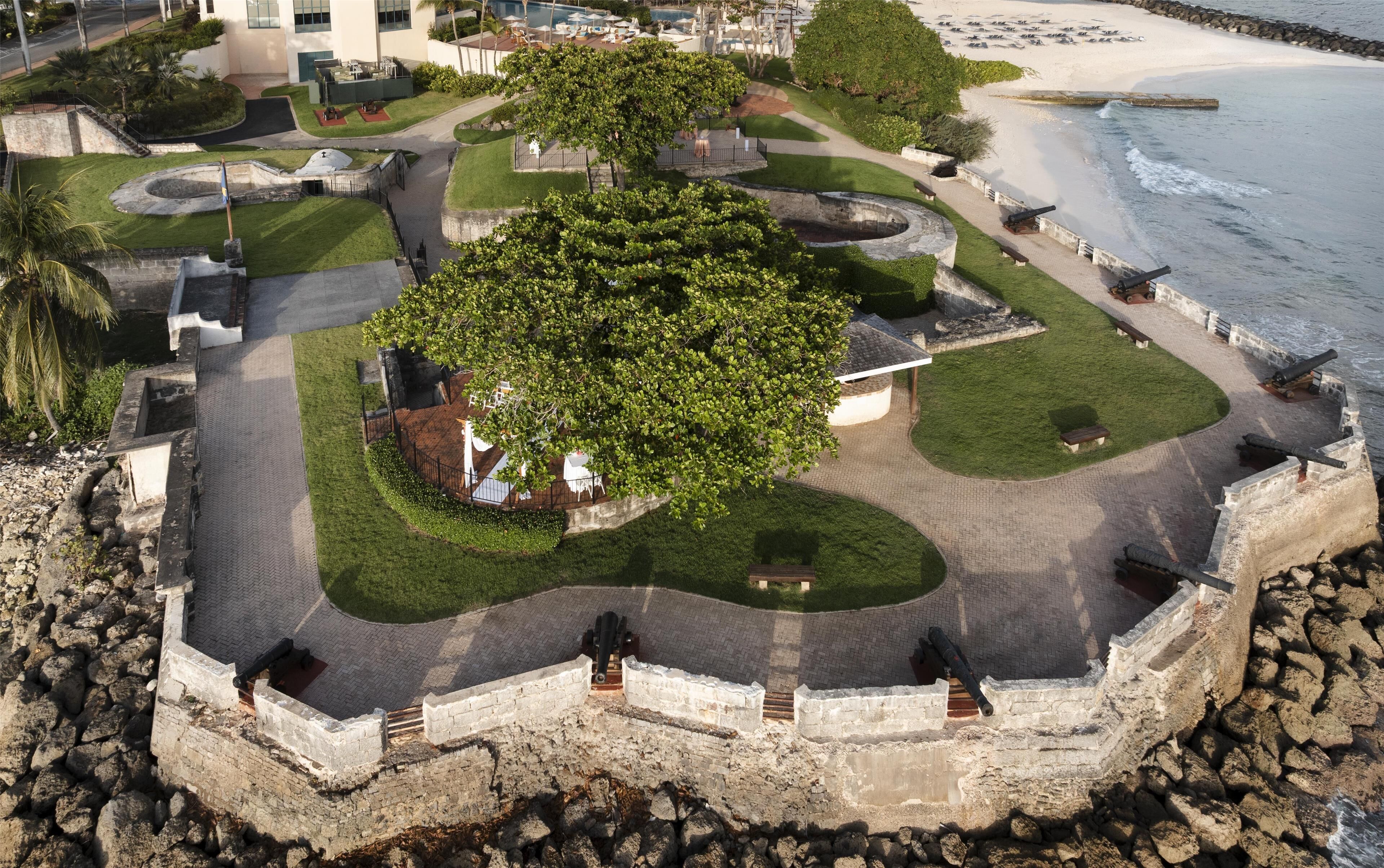 Una fortaleza costera con un árbol central, senderos y una playa al fondo.