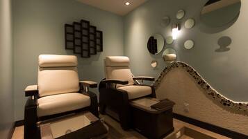 A spa room with two massage chairs, a small table, and a wall decorated with mirrors and a geometric light fixture.