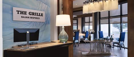 Ein Empfangsbereich mit einem Schild, das 'THE GRILLE BAJAN INSPIRED' zeigt, einer modernen Lampe und Blick auf den Speisesaal mit blauen Stühlen und Holzboden.