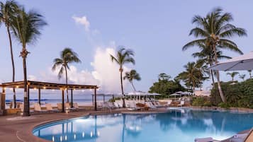 Relax by the serene pool surrounded by palm trees and lounge chairs under a shaded pergola.