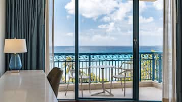 A room with a view of the ocean, a balcony with a table and chairs, and a lamp.