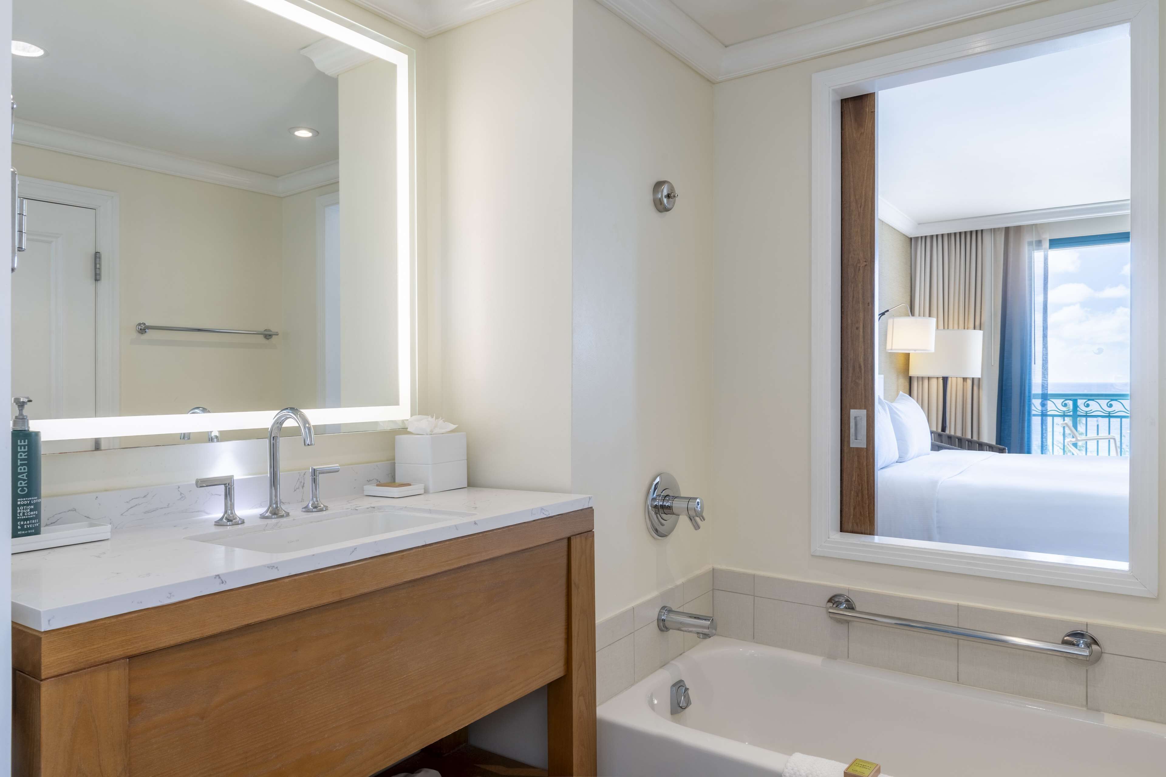 A hotel bathroom with a large mirror, two sinks, a bathtub, and a window with a view of a balcony.