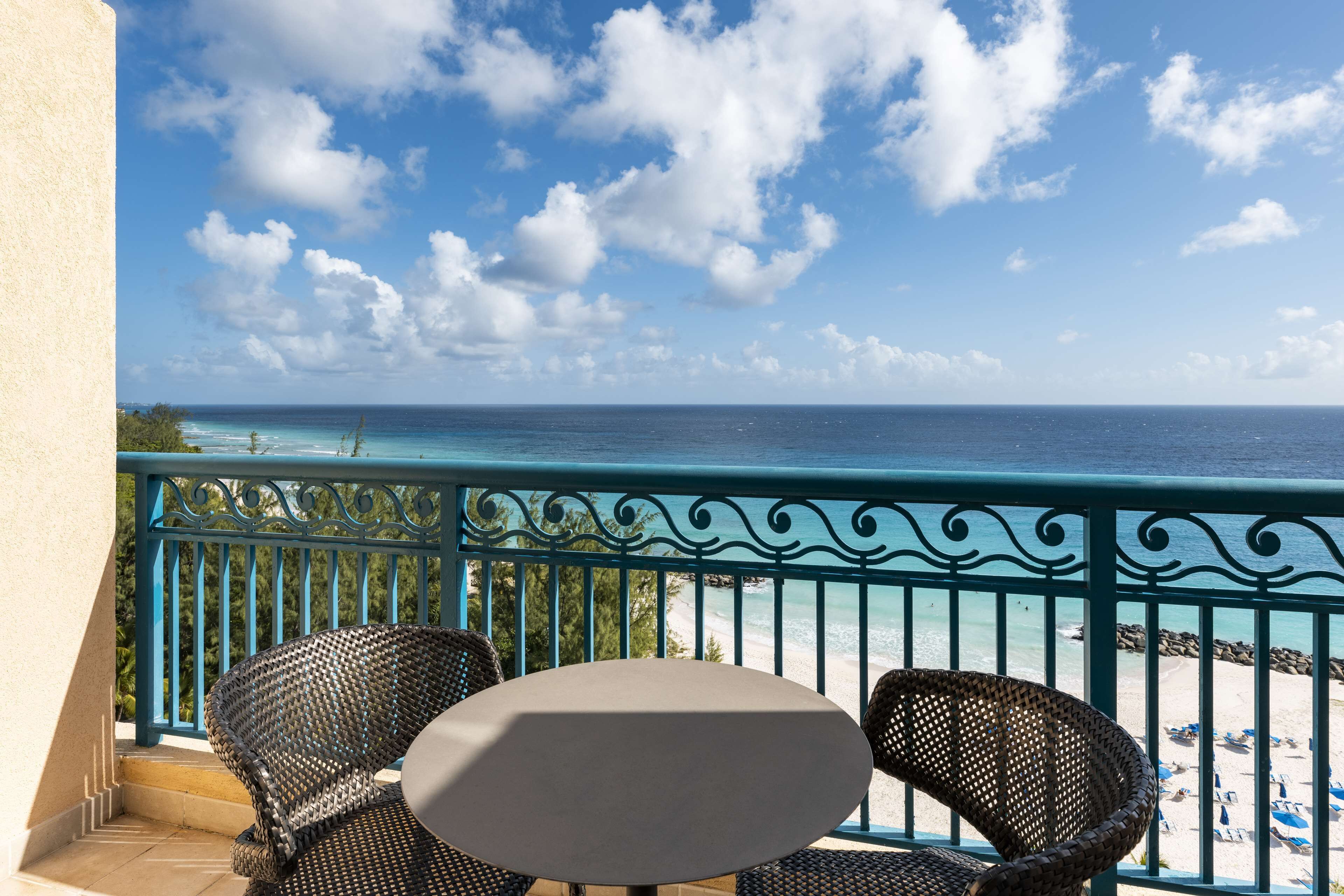 A balcony with wicker chairs and a table overlooking a beach with clear skies.