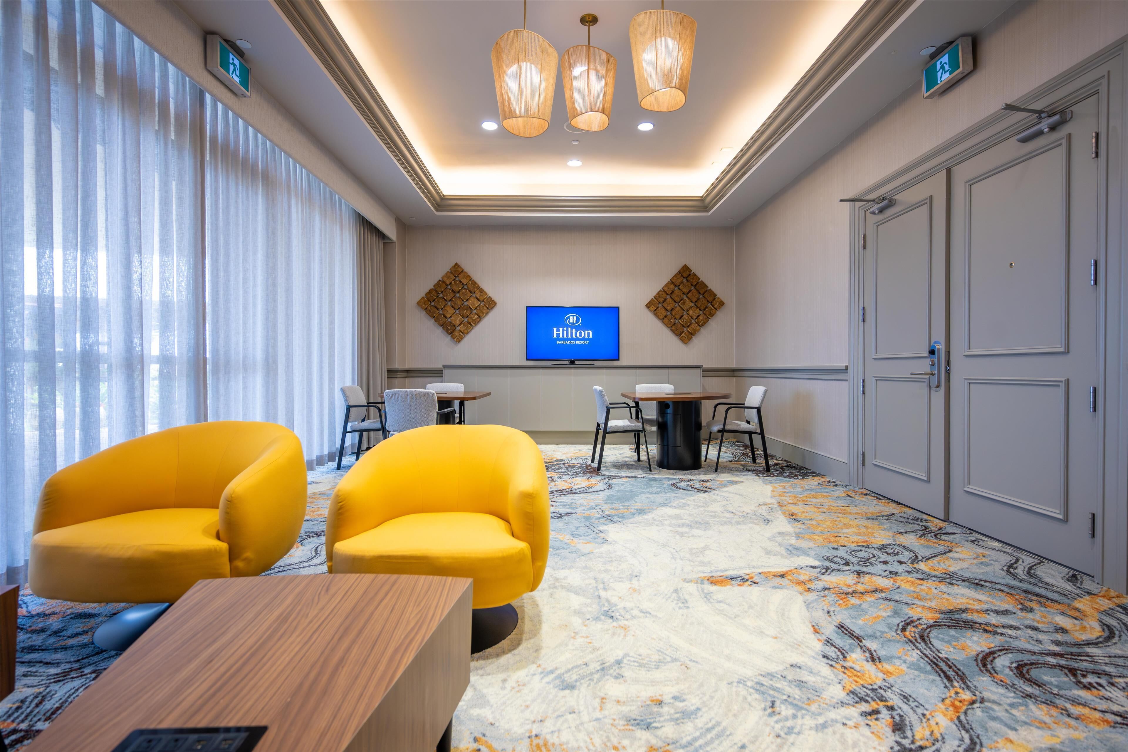 Un vestíbulo de hotel con una pantalla grande mostrando el logotipo de Hilton, dos sillones amarillos, un mostrador de recepción de madera y una alfombra estampada.
