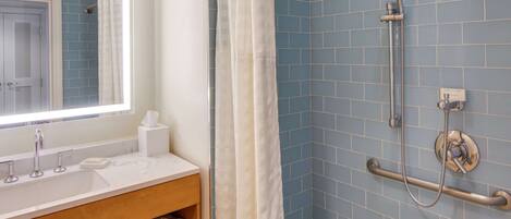 A bathroom with a white sink, a mirror, a shower with a handheld showerhead, and a handrail.
