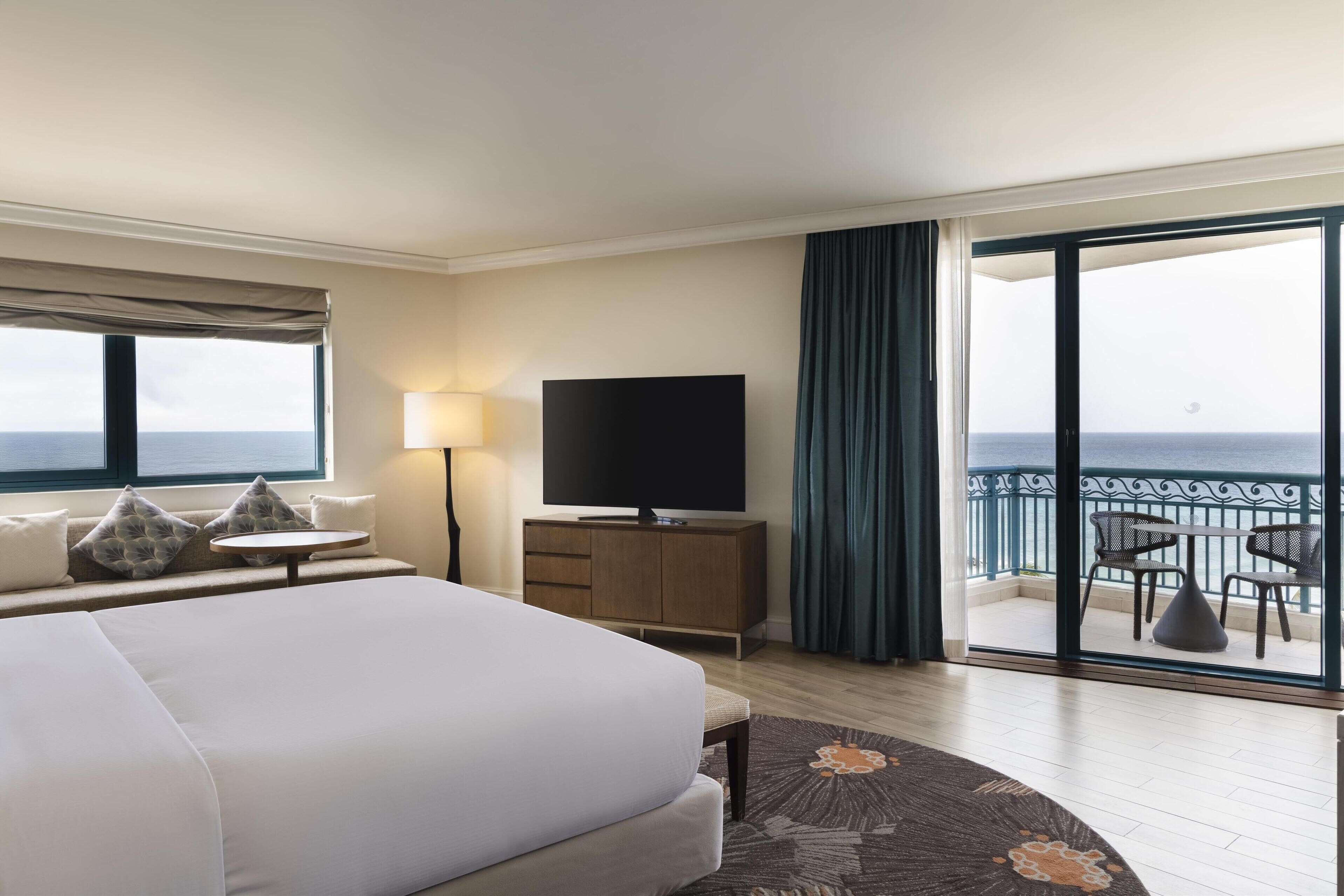A hotel room with a large bed, a flat-screen TV, a bedside table, a lamp, and a balcony with a view of the sea.