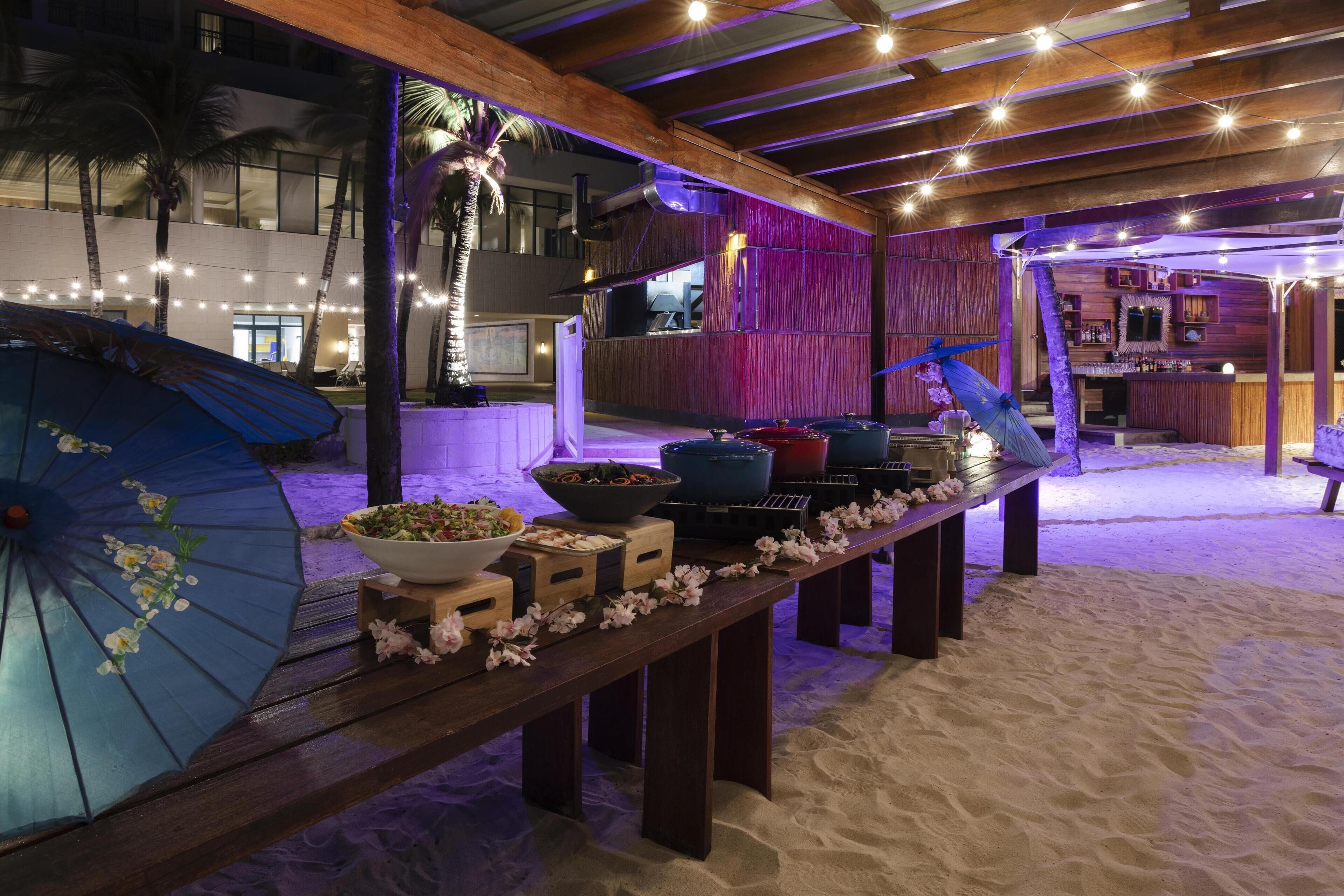 Un área de comedor junto a la playa con una larga mesa de madera dispuesta con comida y bebidas, rodeada de palmeras y luces colgantes.