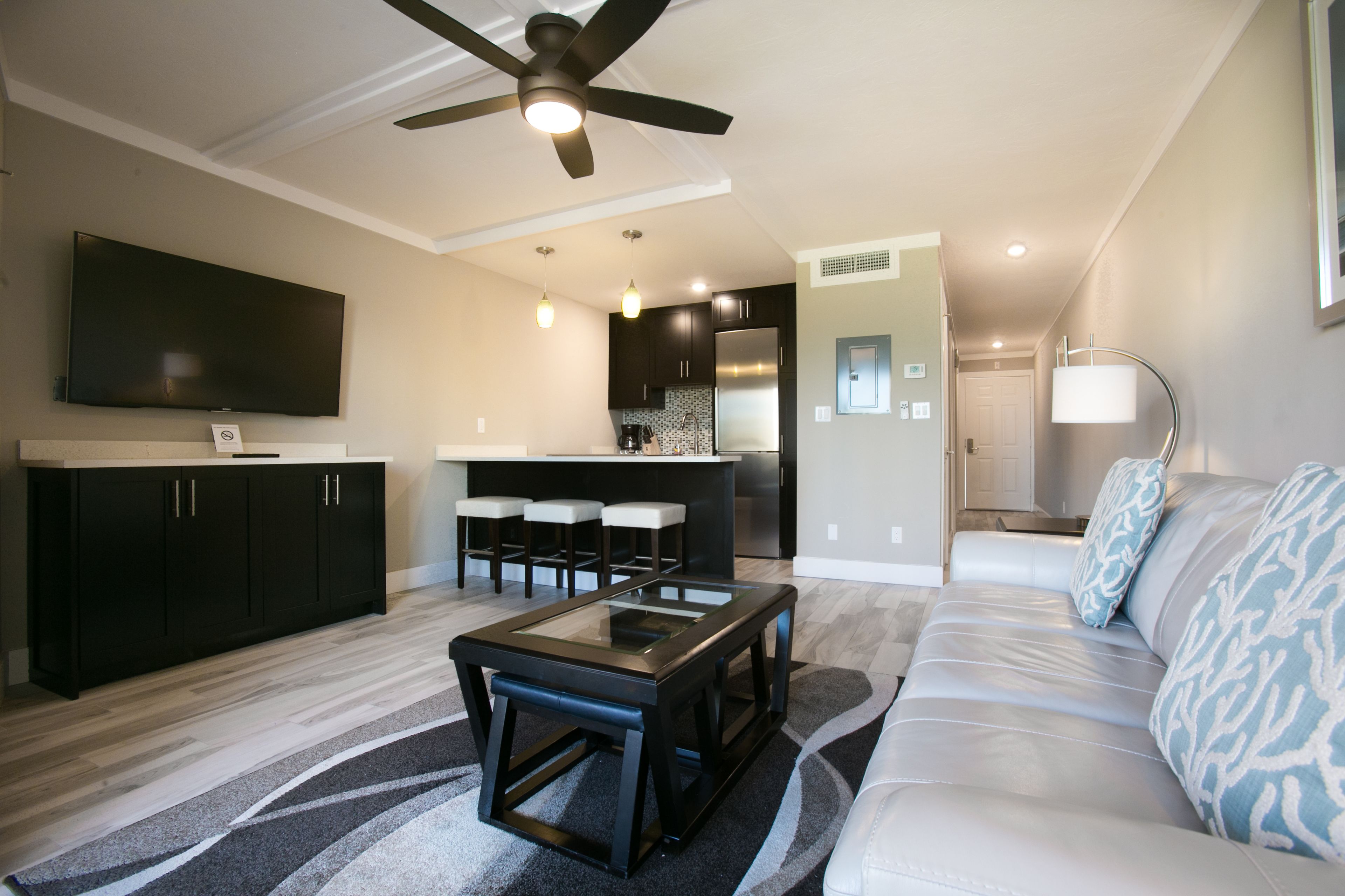 A modern living room with a glass-top coffee table, dark wooden cabinets, a sofa with patterned cushions, a ceiling fan, and a kitchen area in the background.