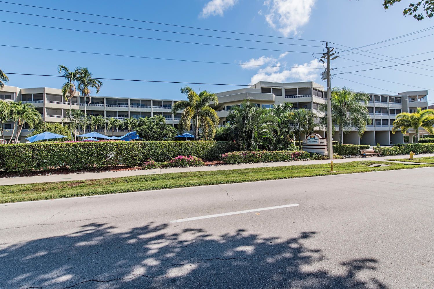 A multi-story hotel with a flat roof, a parking area with blue umbrellas, and a well-maintained garden with palm trees and shrubs.