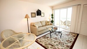 A living room with a sofa, a glass-top table, wicker chairs, a patterned rug, and a balcony with outdoor furniture. - Marco Beach Vacation Suites (Marco Island)