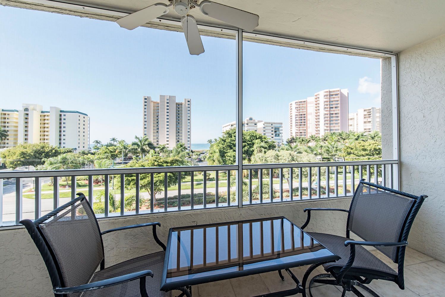 Un balcon avec une table et deux chaises, donnant sur un paysage urbain avec de hauts immeubles et de la verdure.