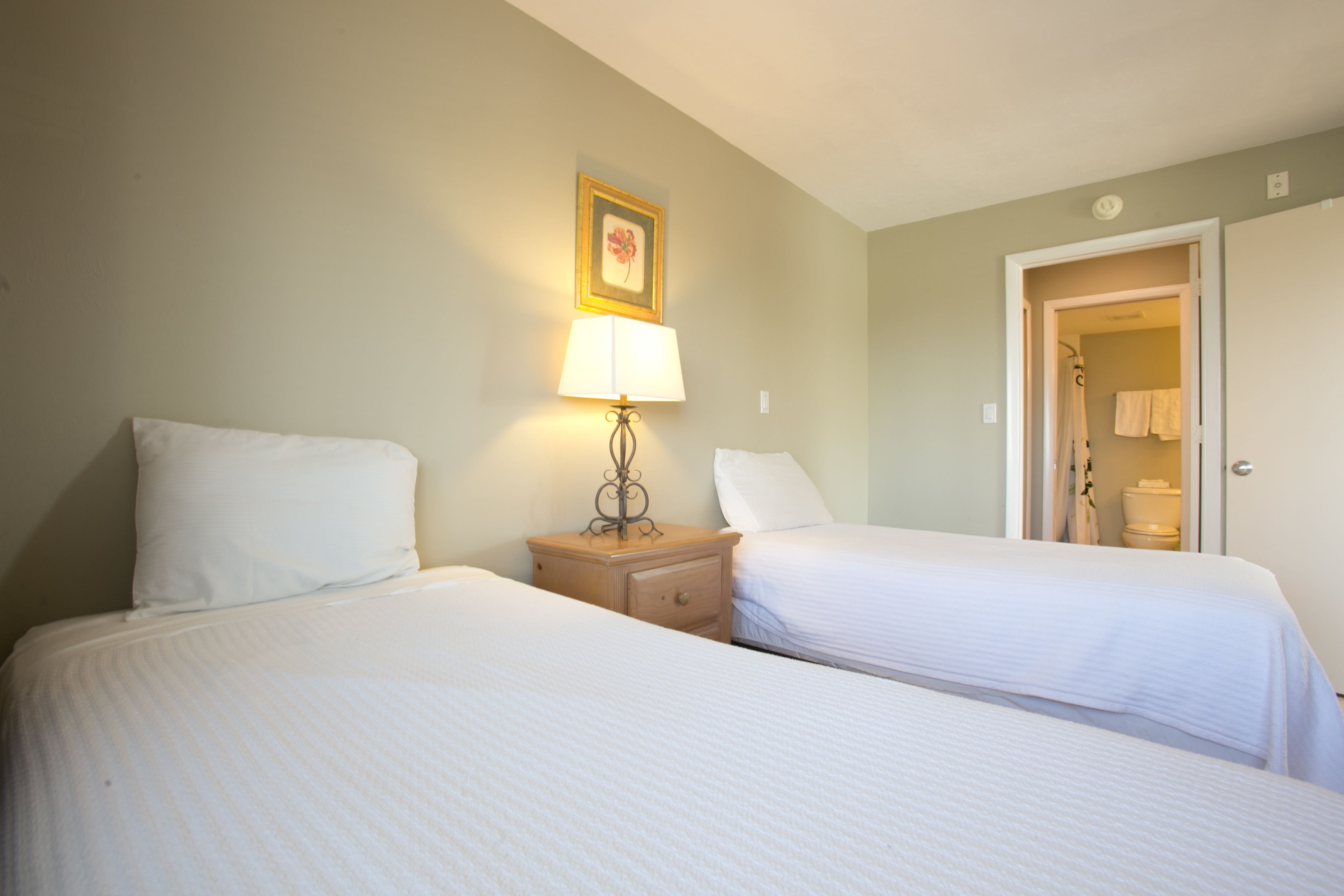 Ein Hotelzimmer mit zwei Betten, einem Nachttisch mit Lampe, einem gerahmten Bild an der Wand und einer Tür zu einem Badezimmer.