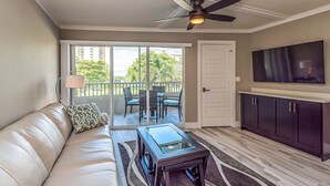 A modern living room with a sofa, a glass coffee table, a TV, and a ceiling fan. - Marco Beach Vacation Suites (Marco Island)