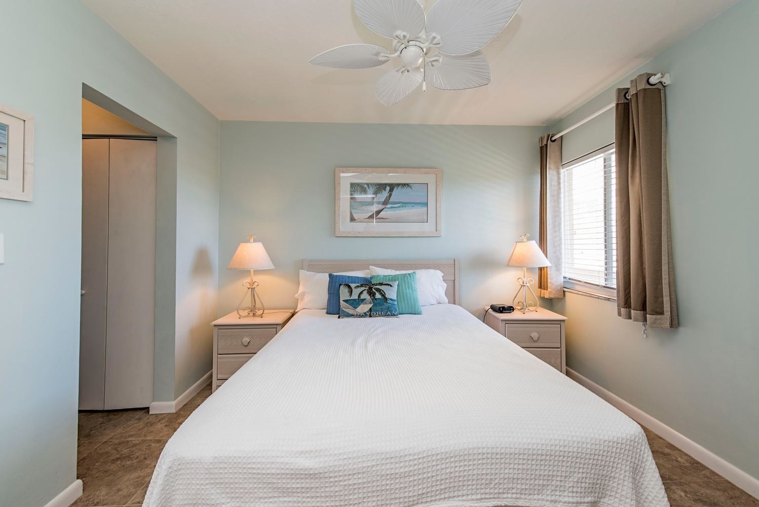 A bedroom with a bed, two bedside tables, a ceiling fan, a window with curtains, and a door.