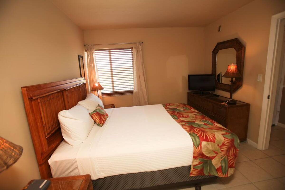 A bedroom with a wooden headboard, a bed with a floral bedspread, a nightstand with a lamp, a dresser, a television, and a mirror on the wall.