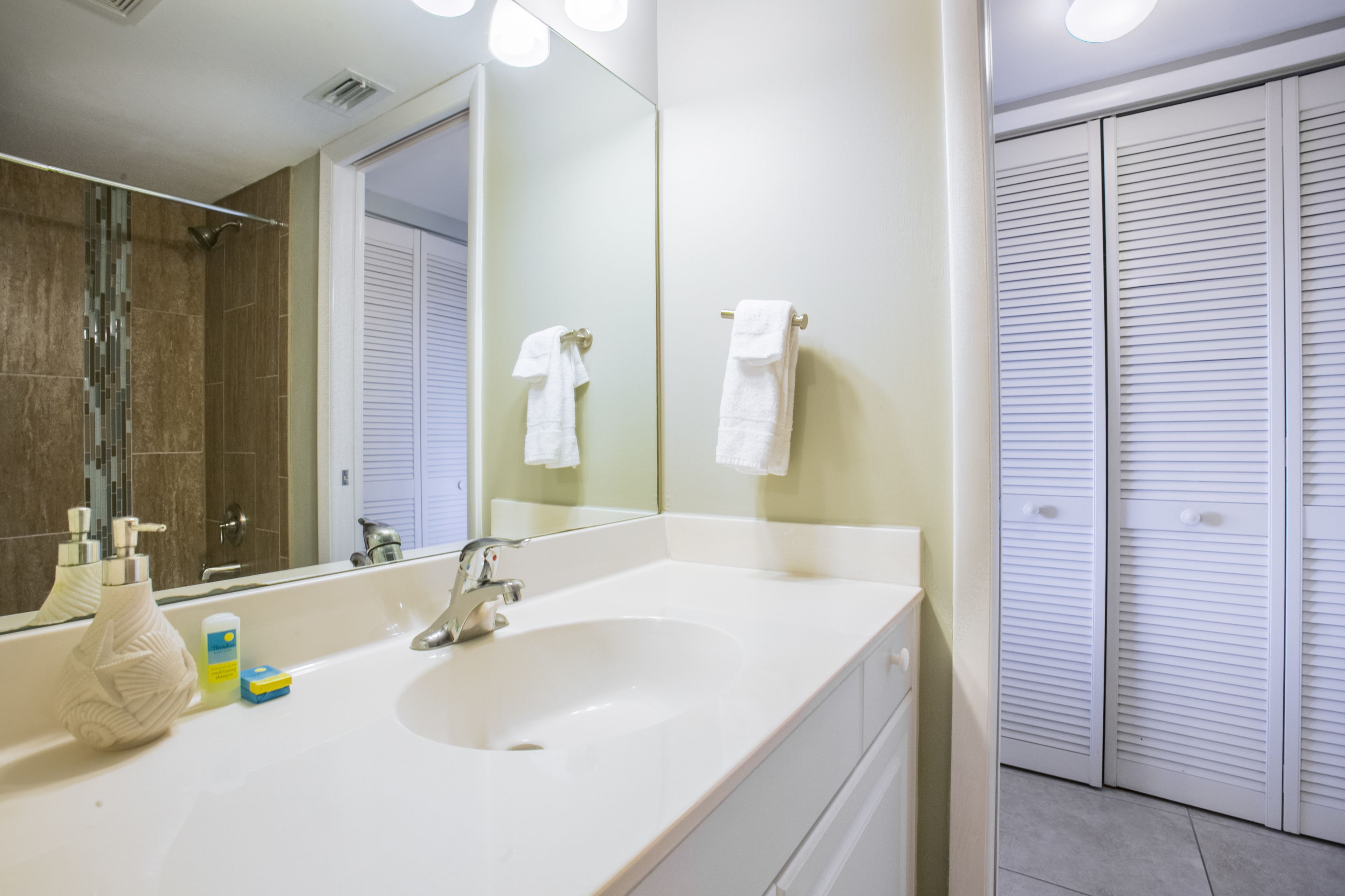 A bathroom with a white sink, a large mirror, and a shower. There are towels hanging on the wall and a toilet visible in the background.