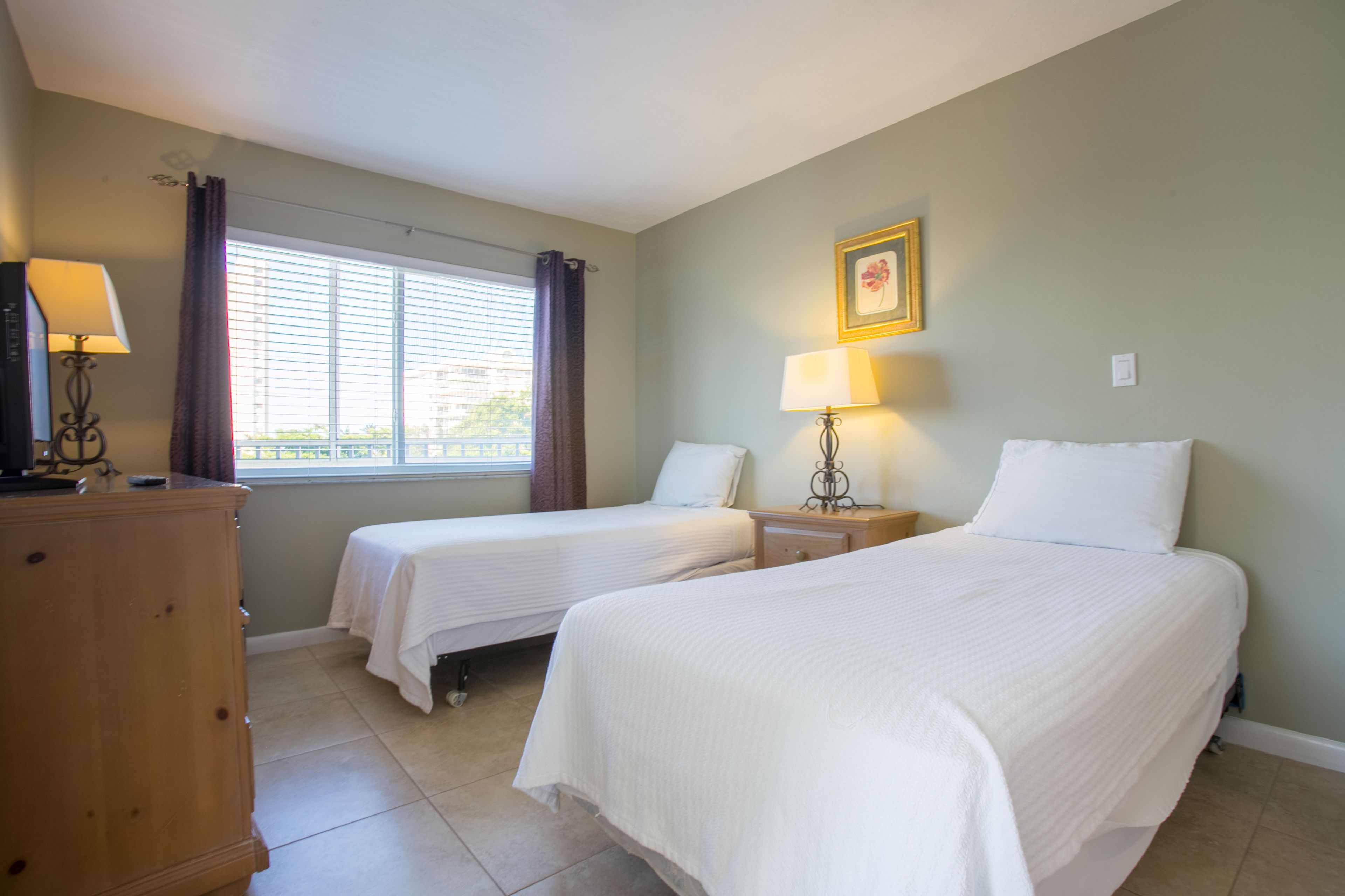 A hotel room with two single beds, a wooden dresser, a window with curtains, and a framed picture on the wall.