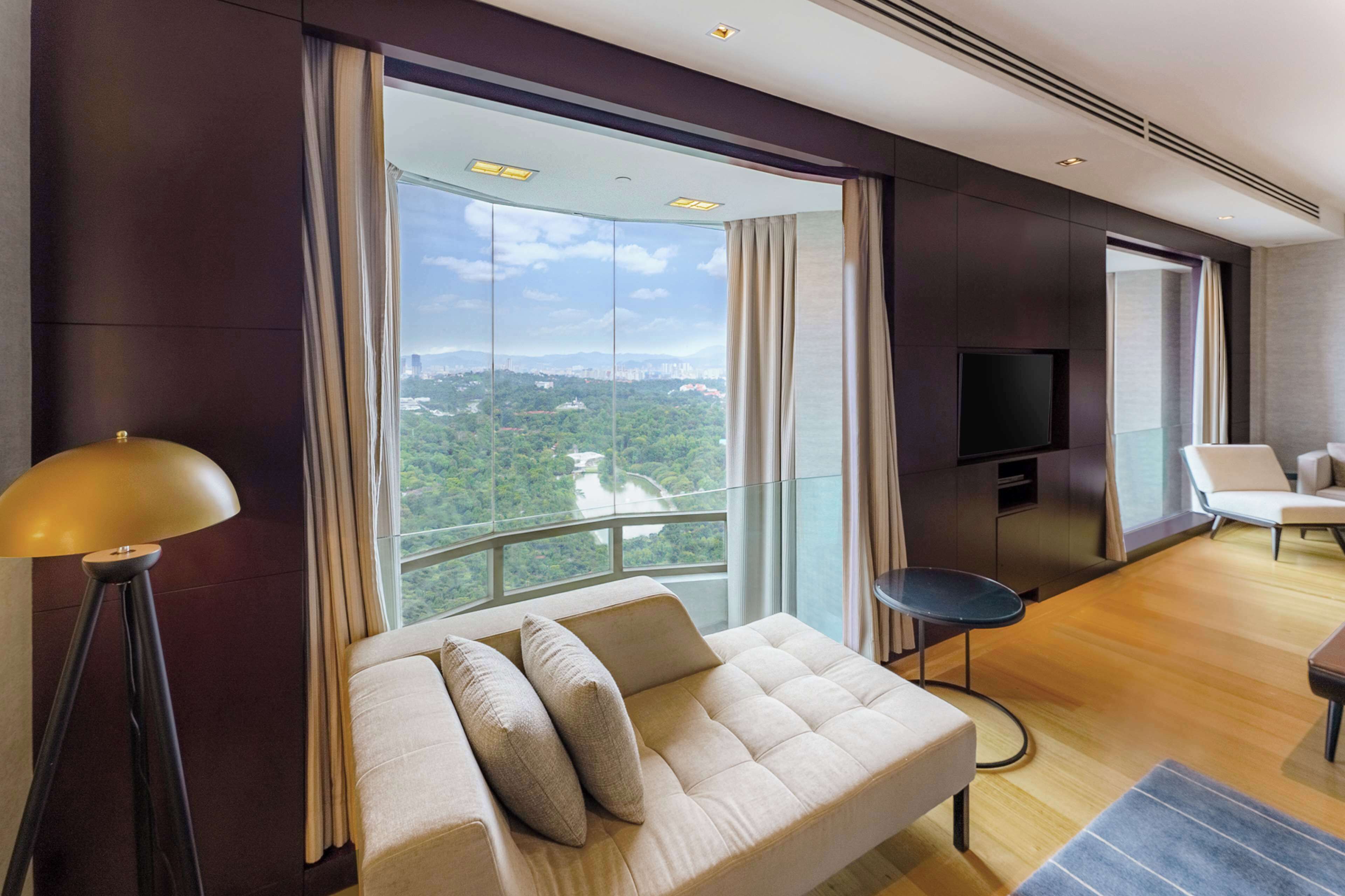 A modern hotel room with a large window offering a city view, a sofa with cushions, a small round table, and a flat-screen TV mounted on the wall.