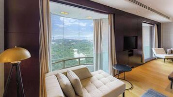 A modern hotel room with a large window offering a city view, a sofa with cushions, a small round table, and a flat-screen TV mounted on the wall.
